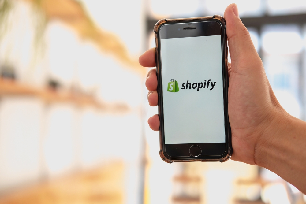 A woman holds iphone 8 plus Mobile Phone with Shopify application on the screen in bakery and coffee shop.